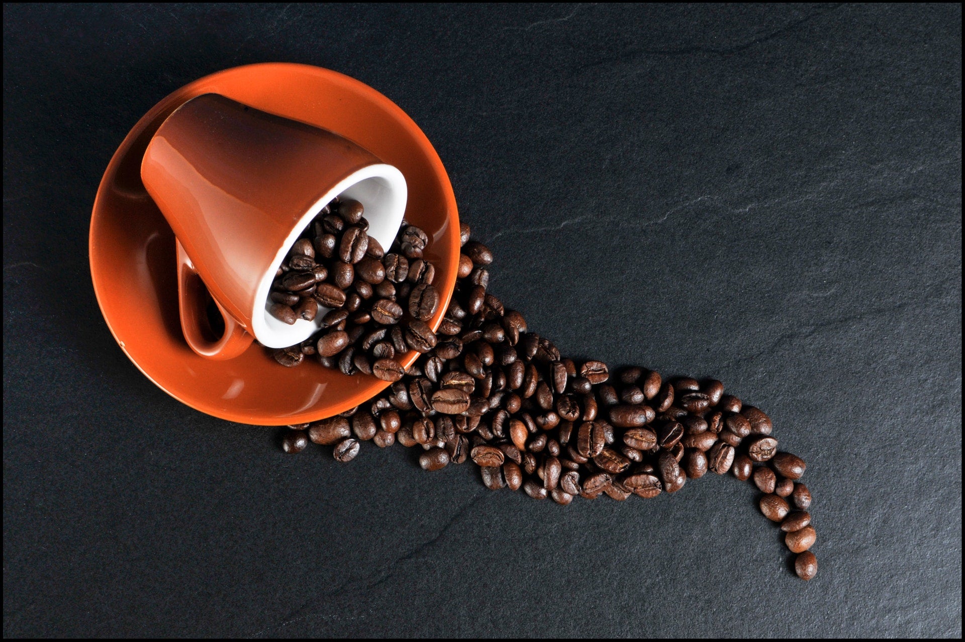 Een omgevallen oranje koffiekopje waaruit geroosterde koffiebonen op een donker oppervlak vallen.